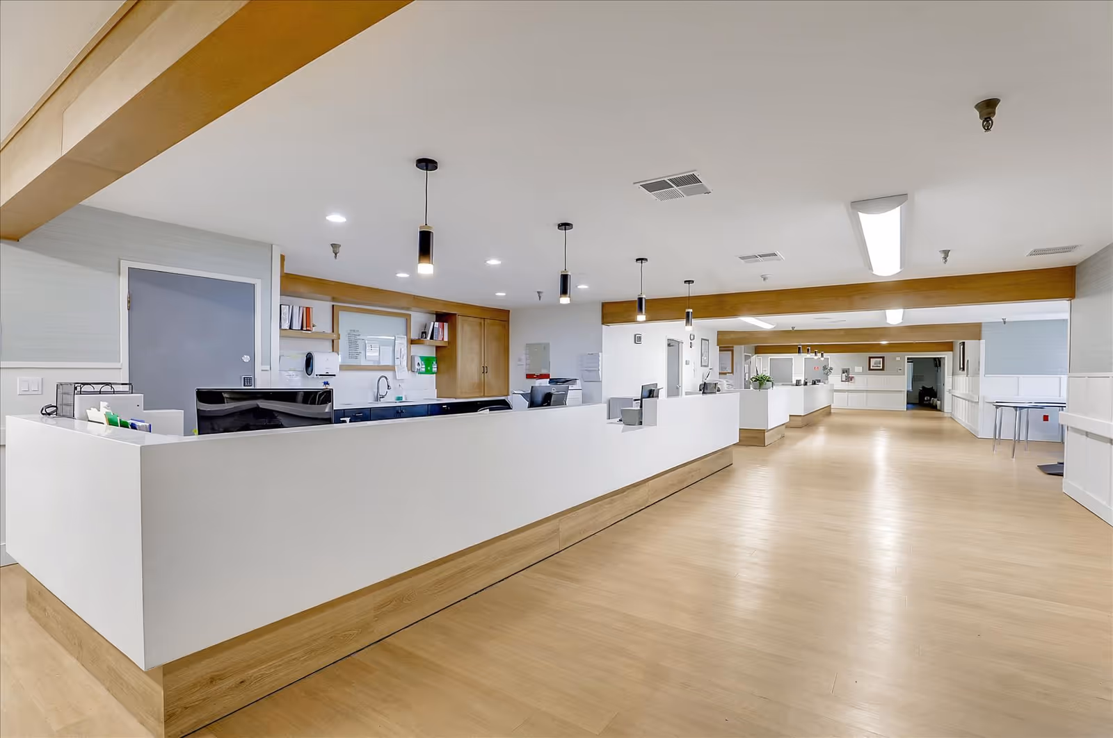 A spacious and well-lit reception or nurse station area in a senior living facility with a long white counter, wooden accents, overhead pendant lights, and a light wood floor. The area is clean and organized with office equipment and bulletin boards visible behind the counter.