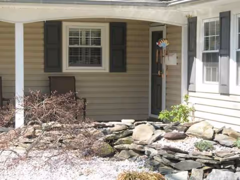 Front entrance of a beige house with shutters, a bench, and rock landscaping.