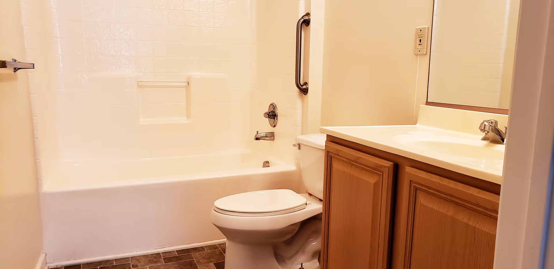 A clean bathroom featuring a white bathtub with a built-in soap holder and a metal grab bar, a white toilet, and a wooden vanity with a sink and faucet. The walls are light-colored, and the floor has dark tiles.