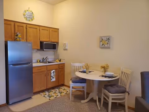 Small kitchen area with wooden cabinets, a stainless steel refrigerator, microwave, and sink. Adjacent to the kitchen is a round dining table set with two chairs, each with a cushion. The table is set with plates, napkins, and decorative items. The walls have simple framed artwork and a decorative sign above the cabinets.
