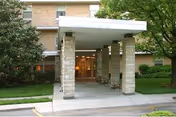 Entrance of a senior living facility named CareCore at Margaret Hall, showing a covered driveway with stone pillars, a concrete walkway, and a brick building facade with windows and greenery on both sides.