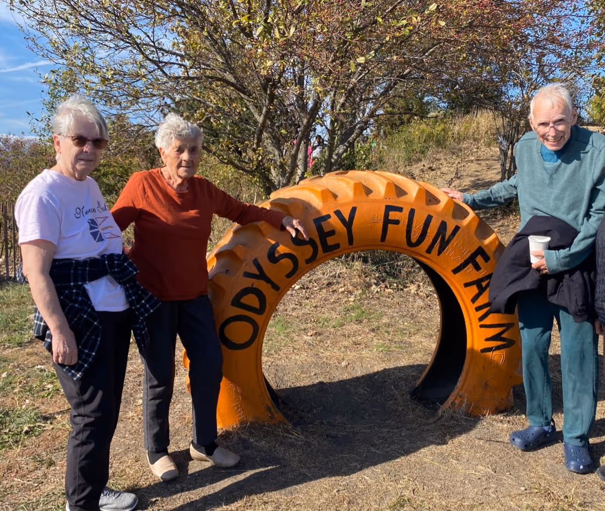 Three elderly individuals standing outdoors next to a large orange tire with the words 'Odyssey Fun Farm' painted on it. They are smiling and appear to be enjoying a sunny day with trees and grass in the background.