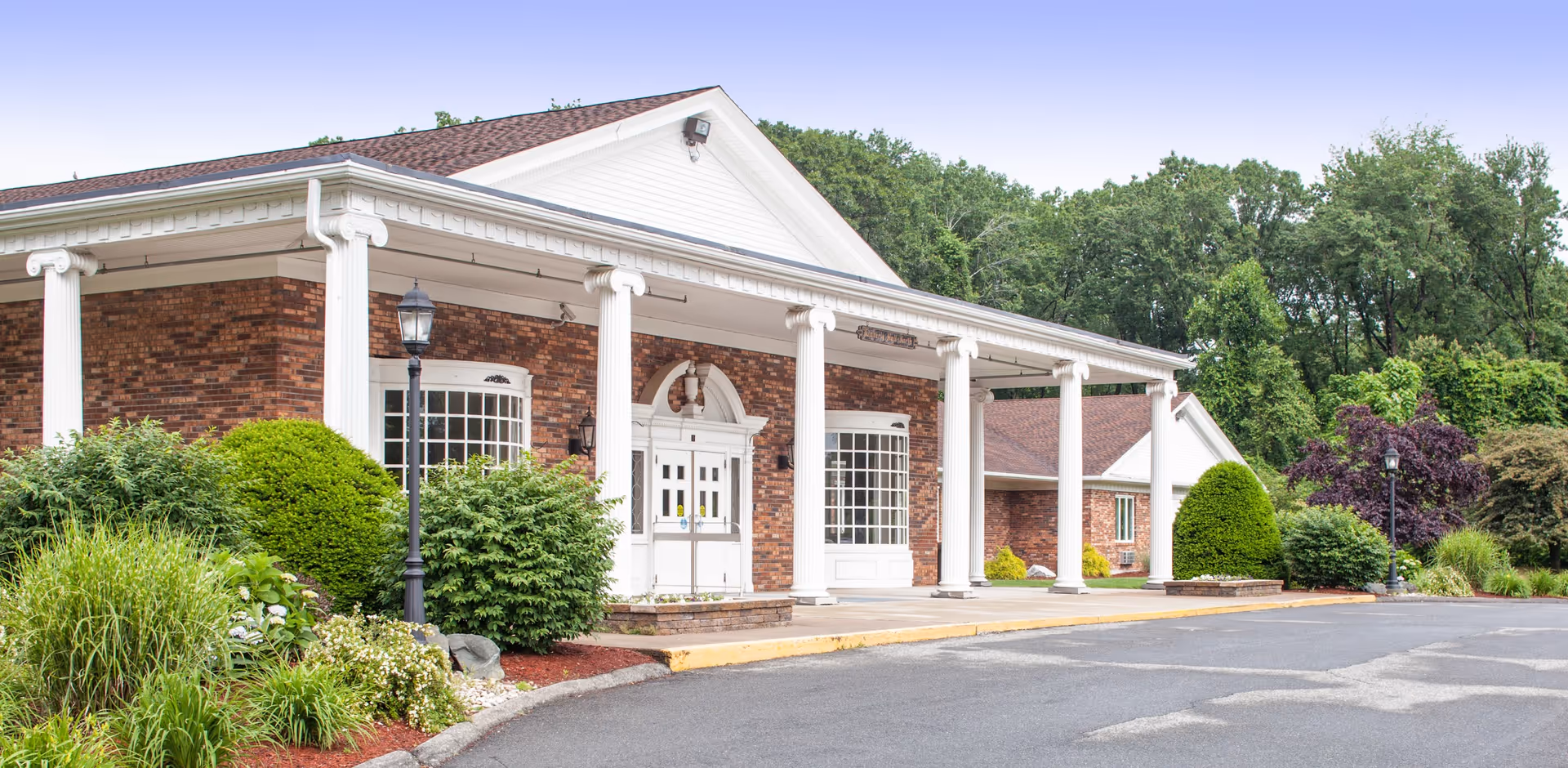 Front exterior view of a brick building with white columns and a white entrance door, surrounded by green bushes and trees under a clear sky.