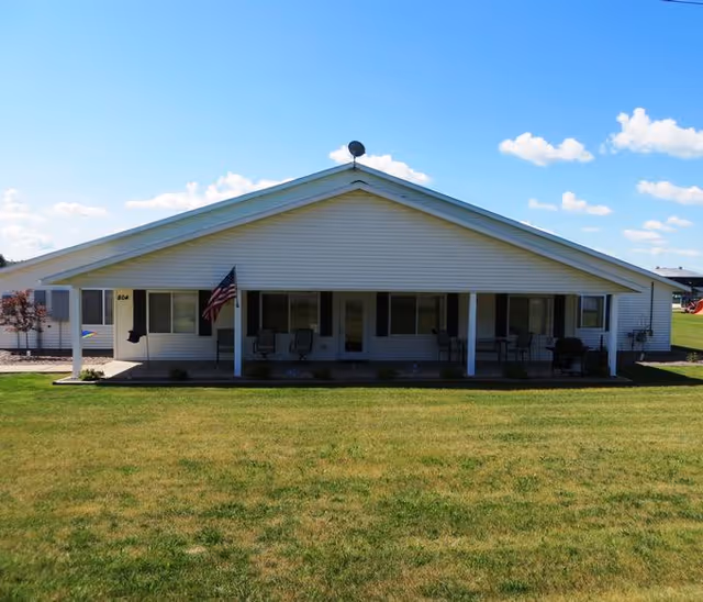 Single-story white ranch-style building with a covered front porch, chairs and an American flag in front of a grassy lawn.