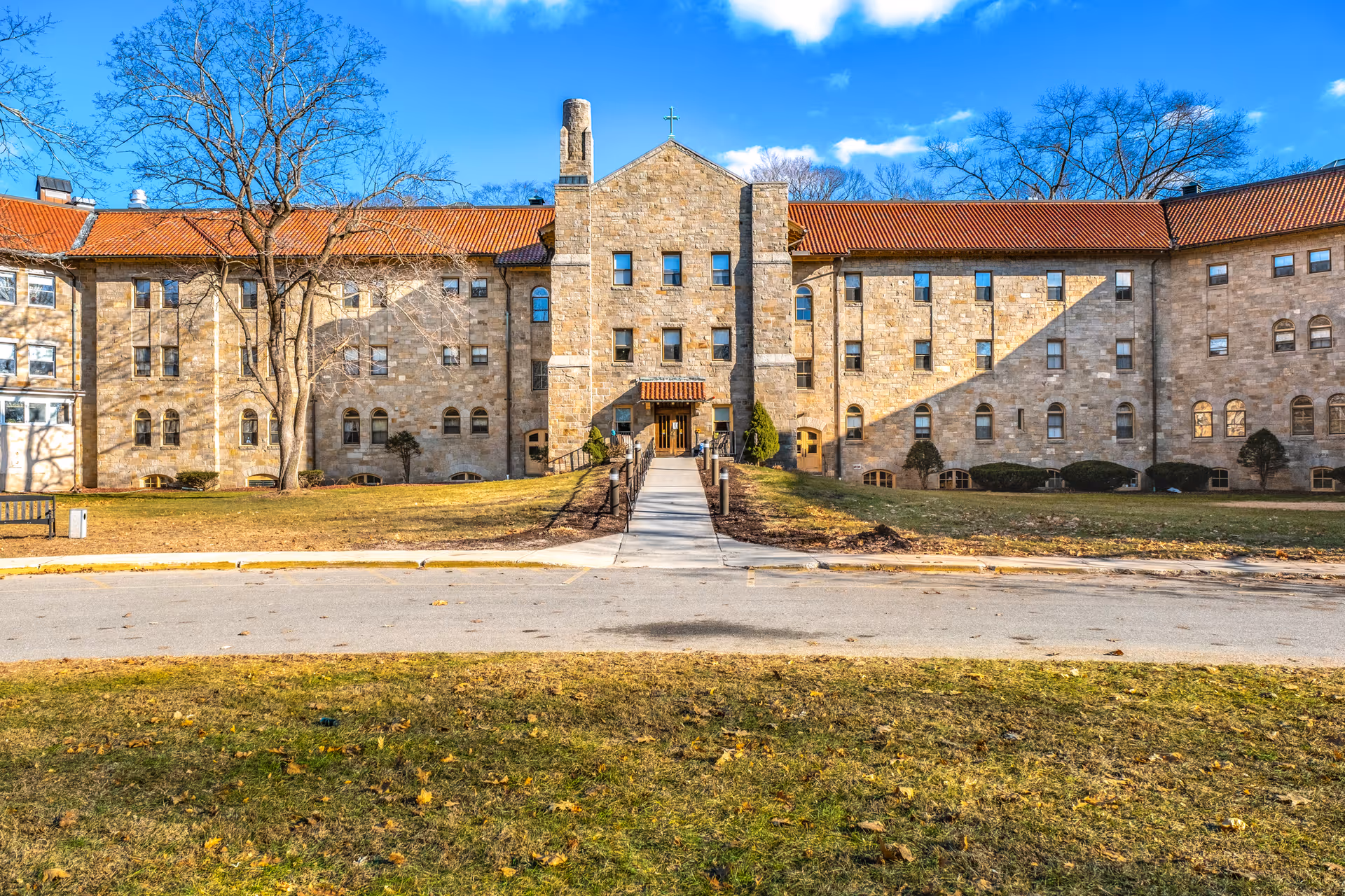 Front exterior view of a large stone building with a red tile roof, multiple windows, and a central entrance with a small awning. There is a paved walkway leading up to the entrance, surrounded by grass and a few trees without leaves. The sky is clear and blue.