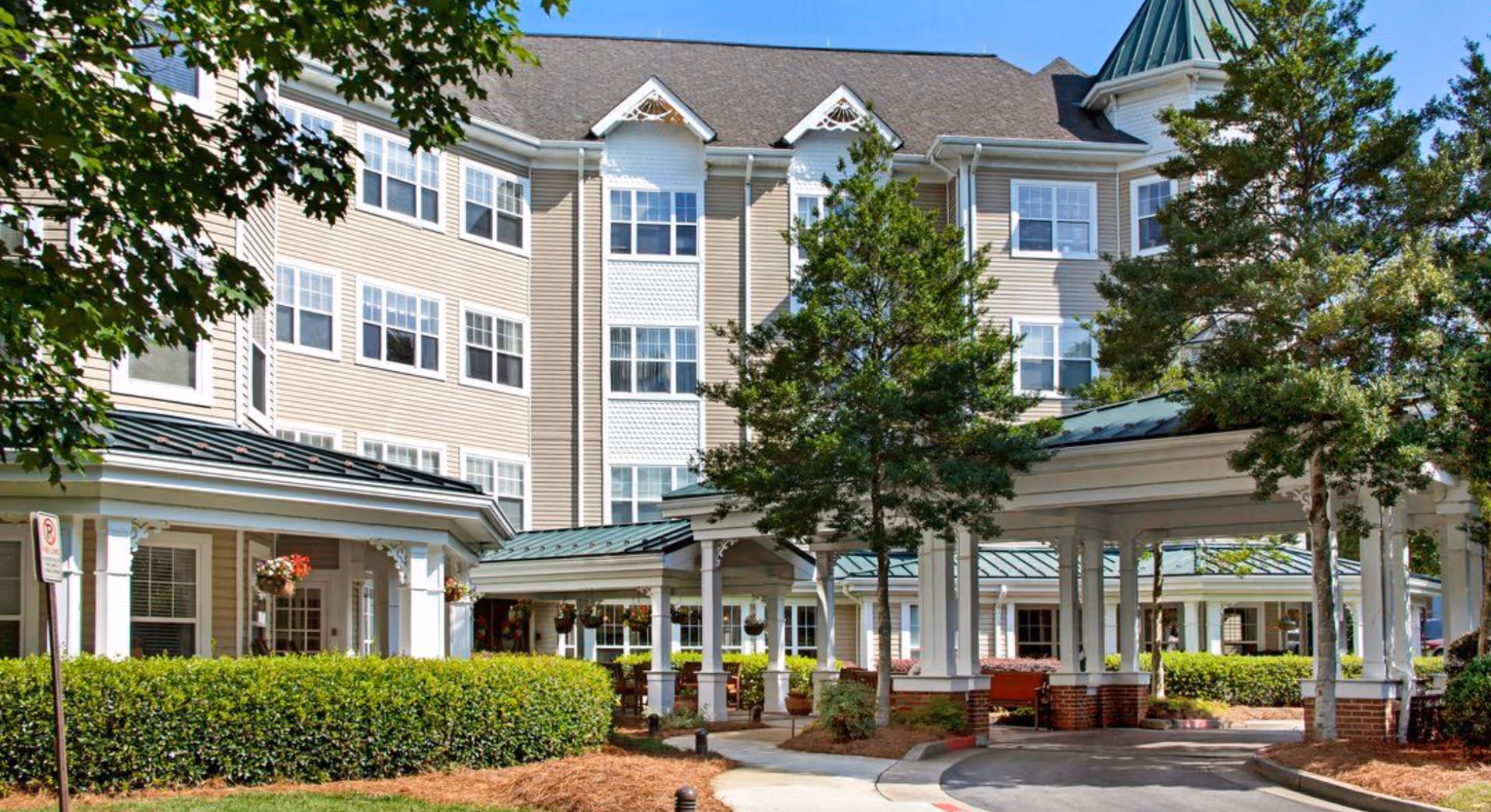 Front exterior of a multi-story senior living building with a covered porte-cochere, manicured hedges, and trees.