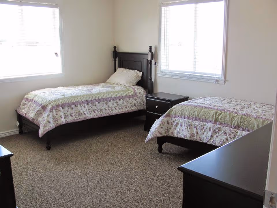 A bright bedroom with two single beds featuring floral and striped bedspreads in shades of purple and green. Each bed has a dark wooden headboard and there is a matching dark wooden nightstand with two drawers between the beds. The room has beige carpet flooring and two windows with white blinds allowing natural light to enter.