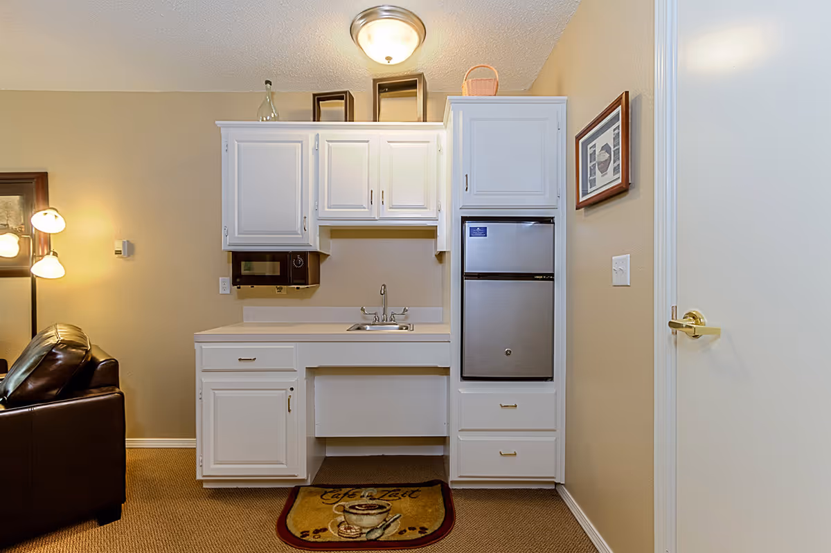 A small kitchenette area with white cabinets, a stainless steel mini refrigerator, a microwave, a sink, and a countertop. To the left, part of a brown leather chair and a floor lamp with three lights are visible. The walls are beige, and there is a framed picture on the right wall. A small rug with a coffee cup design is placed on the floor in front of the kitchenette.