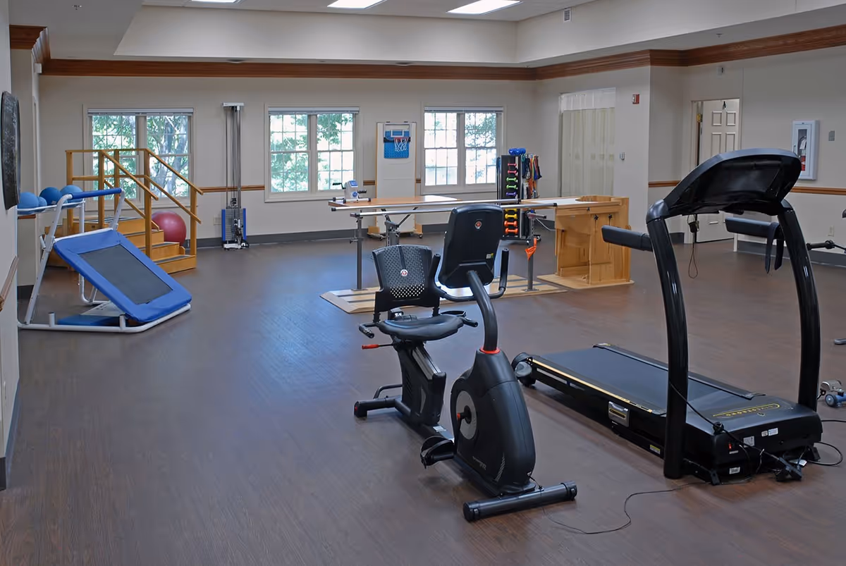 A spacious senior living facility exercise room with various fitness equipment including a recumbent exercise bike, treadmill, small trampoline, parallel bars for walking support, and a rack of colorful dumbbells. The room has large windows letting in natural light and wooden trim along the ceiling and walls.