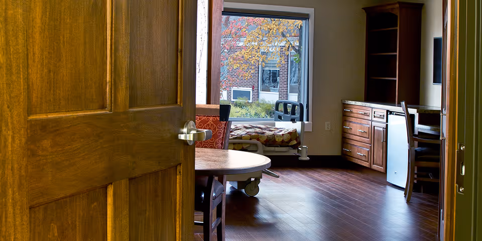 Partially open wooden door reveals a furnished studio room with a round table, a hospital-style bed on wheels, cabinets, and a window.