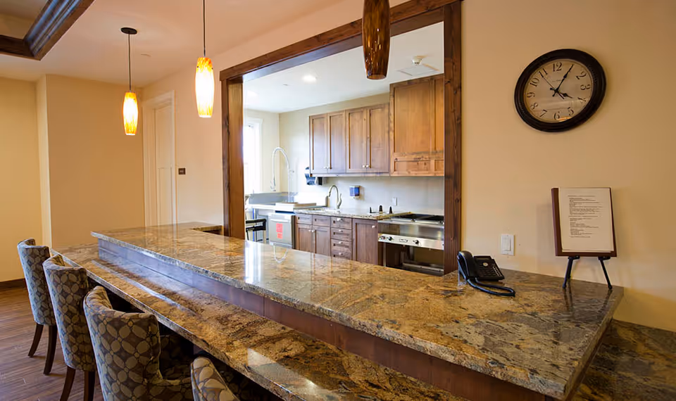 Granite-topped serving counter with barstools and pendant lights overlooking a kitchen with wooden cabinets and a wall clock.