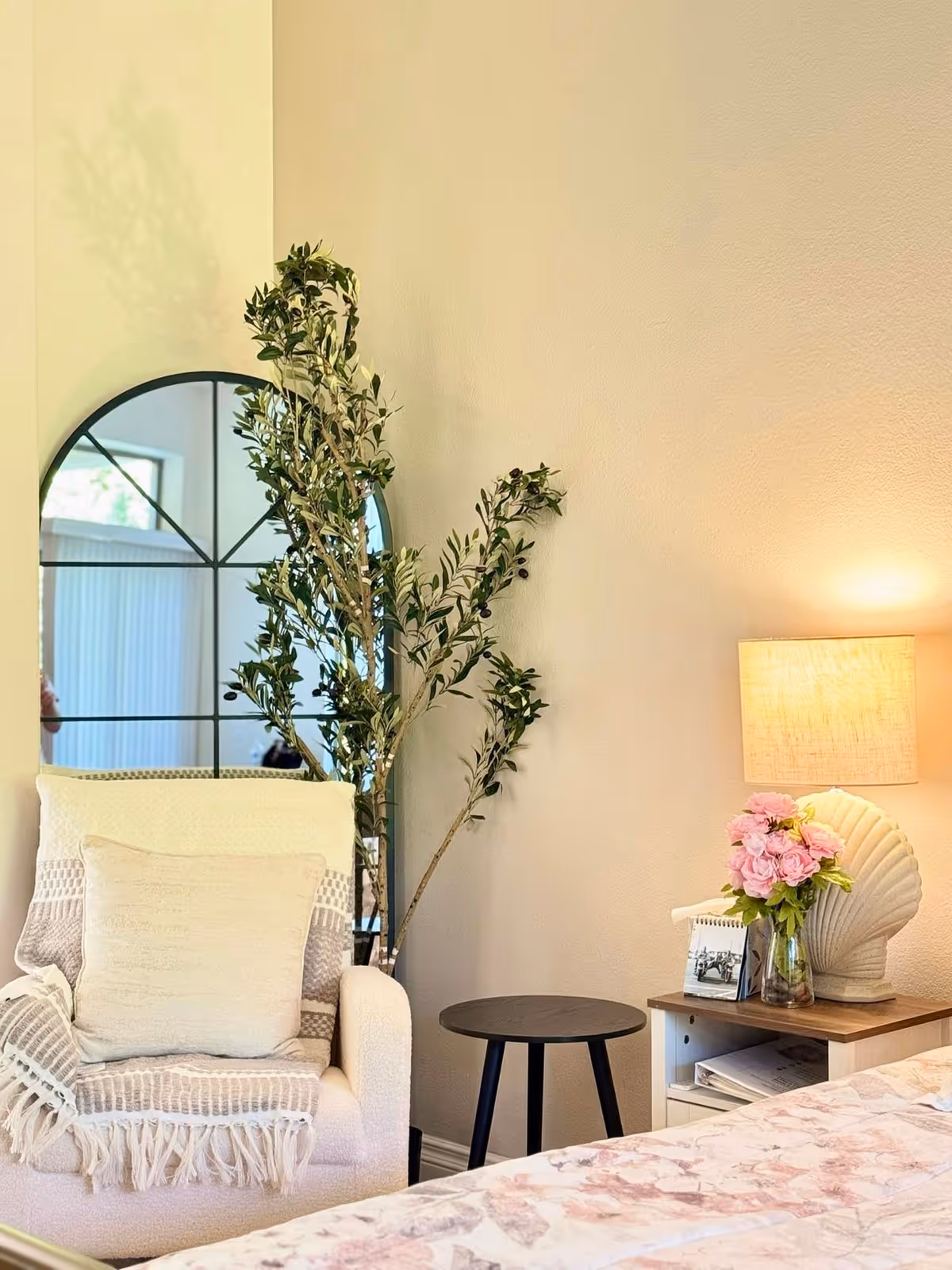 Cozy corner of a bedroom featuring a white armchair with a textured throw and pillow, a tall leafy plant behind it, a round black side table, and a bedside table with a shell-shaped lamp, a vase of pink flowers, and a small photo frame. The wall is painted light beige and a large window is reflected in a decorative arched mirror behind the plant.