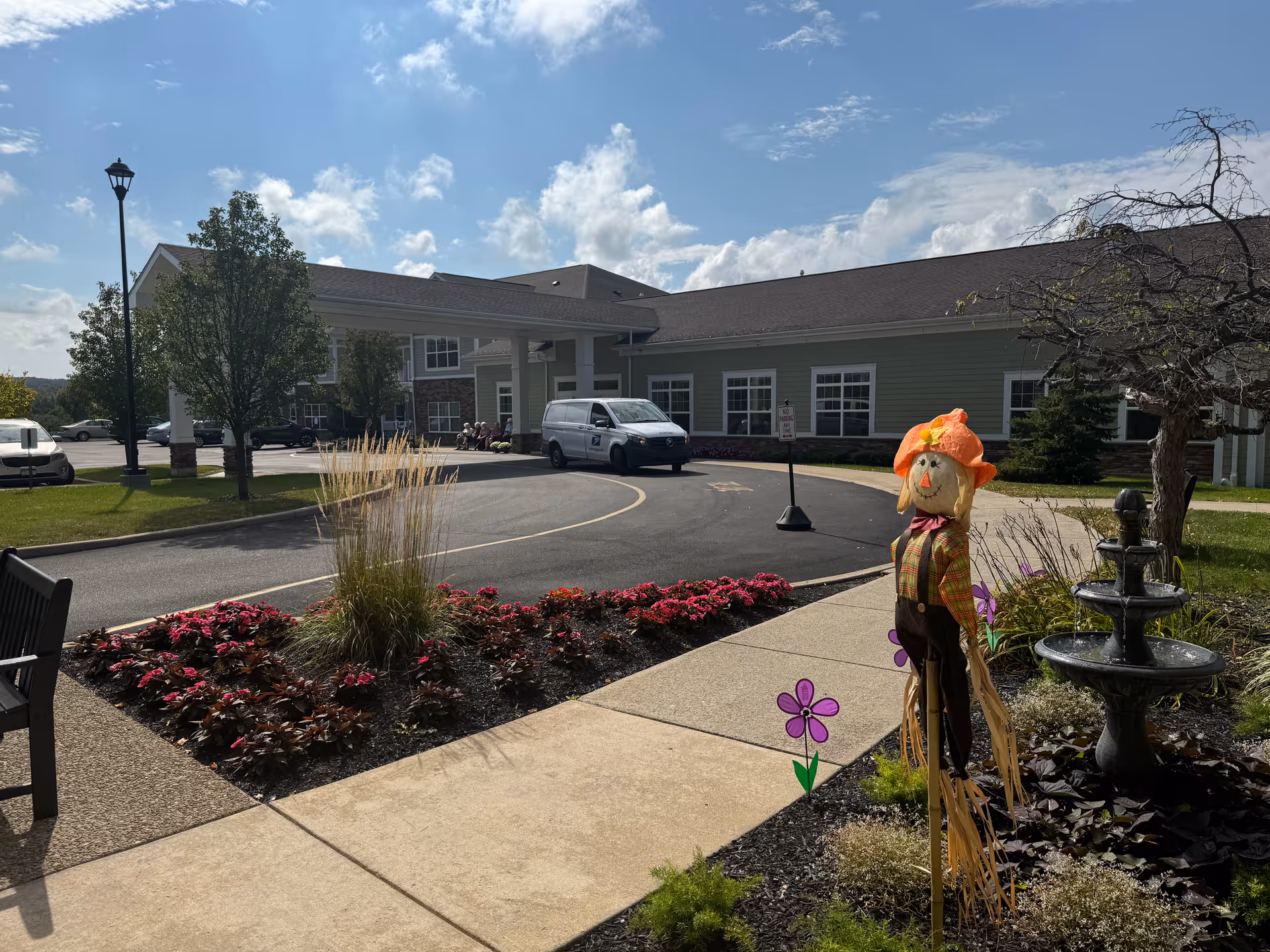 Outdoor view of Orchard Heights facility in Orchard Park showing a paved driveway, landscaped flower beds with pink flowers, a scarecrow decoration, a black water fountain, a bench, and a white van parked near the building entrance under a covered drop-off area. Trees and a lamppost are also visible under a partly cloudy sky.