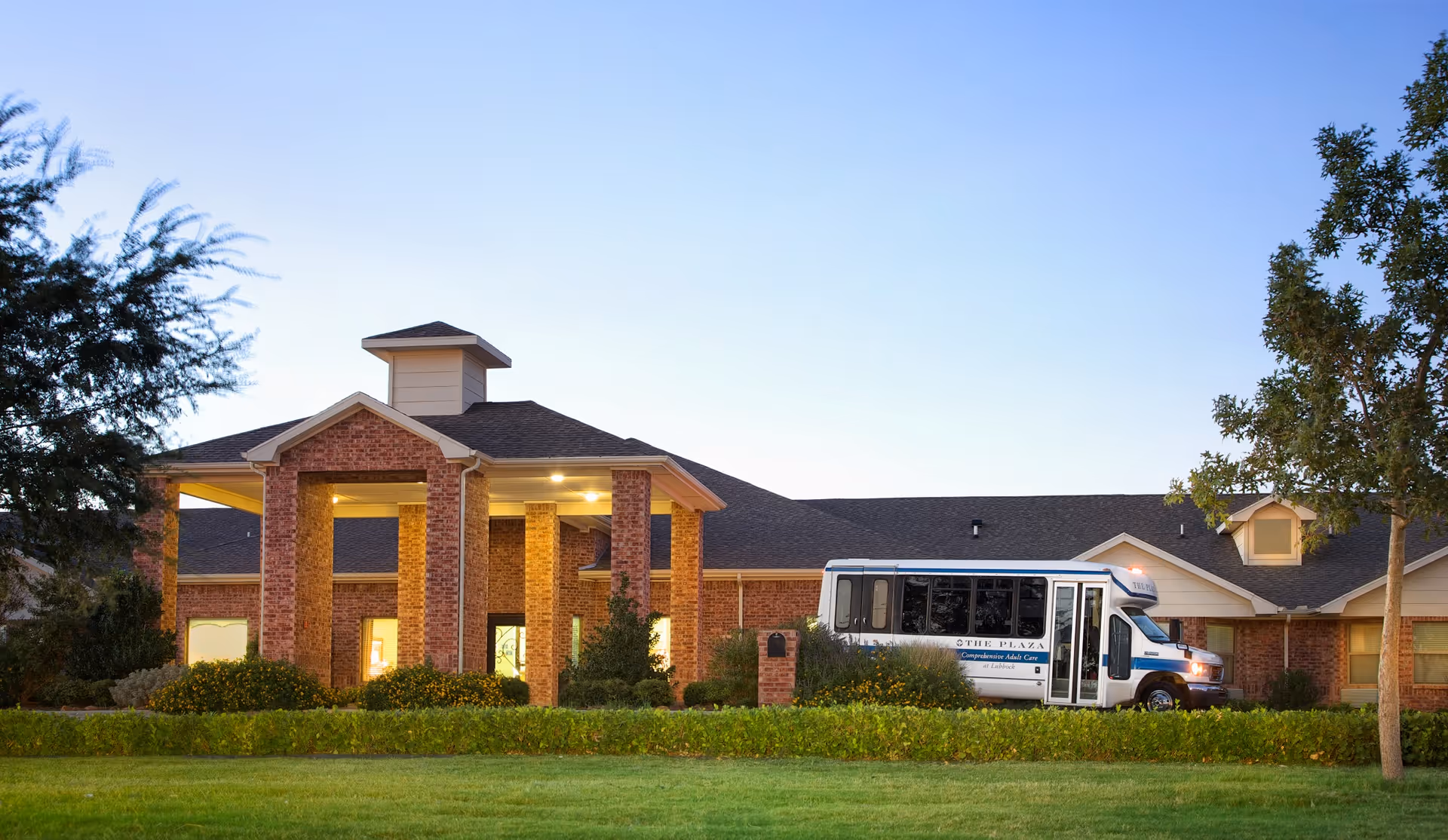 Exterior view of The Plaza At Lubbock facility during dusk, showing a brick building with a covered entrance and a white shuttle bus parked in front. The surrounding area includes green grass, bushes, and trees under a clear sky.