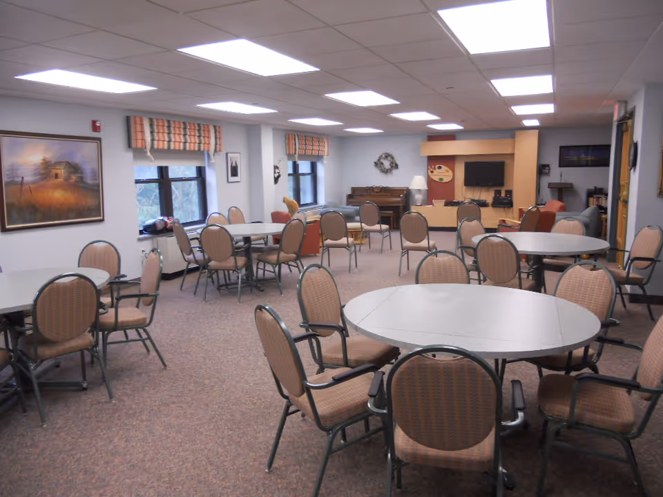 A spacious room with multiple round and rectangular tables surrounded by chairs. The room has carpeted floors, several windows with patterned valances, a piano, a television mounted on a wooden cabinet, and various wall decorations including paintings and a wreath.