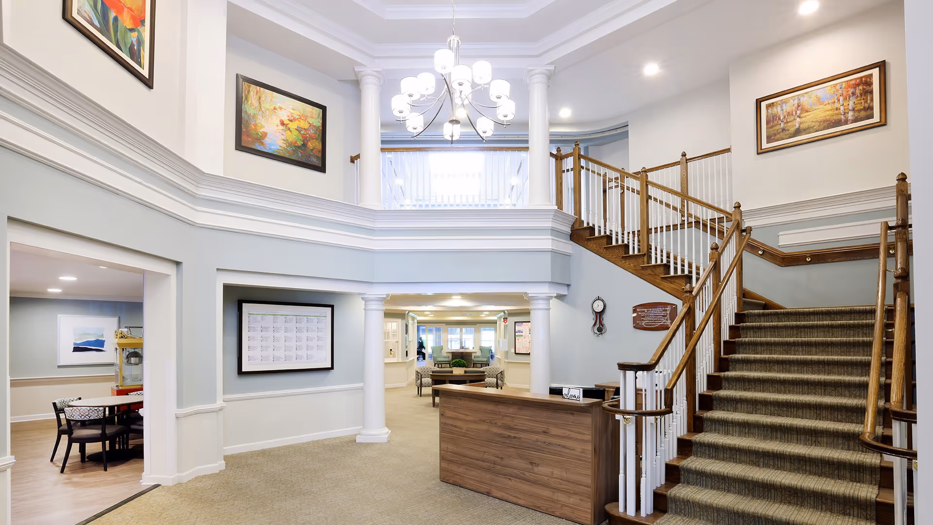 Interior view of a senior living facility lobby area with a wooden reception desk in front of a carpeted staircase with wooden handrails. The space features white columns, light blue and white walls, framed artwork, and a chandelier hanging from the ceiling. There is a seating area visible in the background and a dining area with tables and chairs to the left.