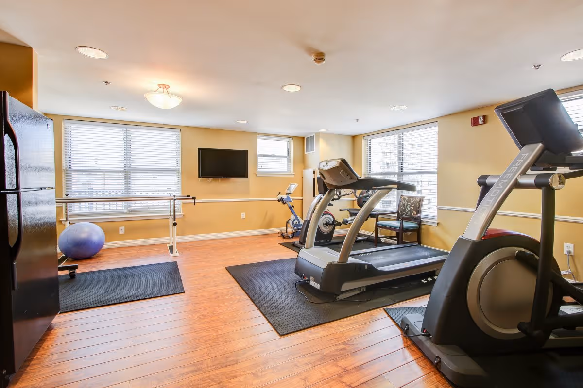 A well-lit fitness room with wooden floors and yellow walls featuring two treadmills, an exercise bike, a large blue exercise ball, a wall-mounted TV, and large windows with blinds allowing natural light to enter.