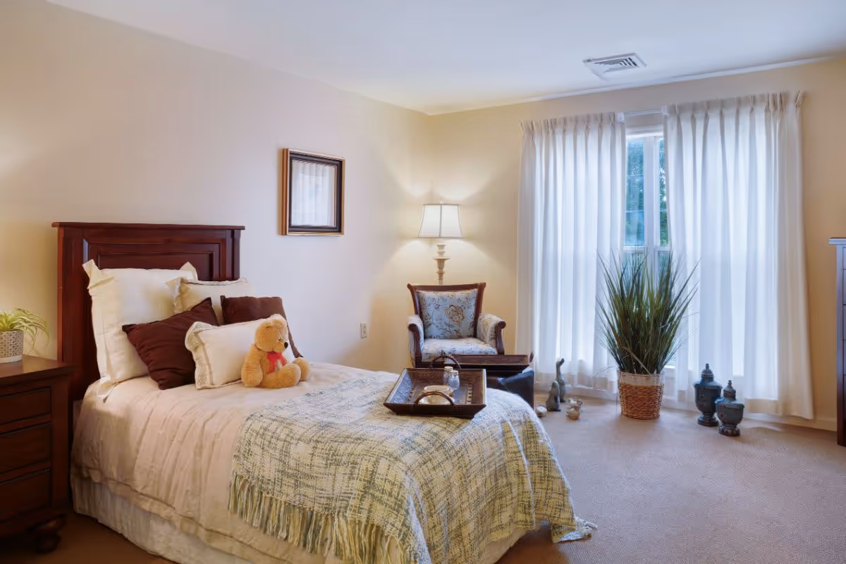 A cozy bedroom with a single bed adorned with multiple pillows and a teddy bear. Next to the bed is a wooden nightstand with a small plant. A cushioned armchair with a floral pattern sits near a window with sheer white curtains. A floor lamp provides warm lighting, and decorative items including a potted plant and small statues are placed near the window.