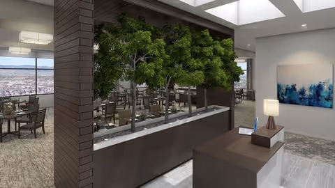 Interior view of a senior living facility showing a modern reception desk with a lamp and decorative items, a planter with green trees, and a dining area with tables and chairs in the background near large windows.