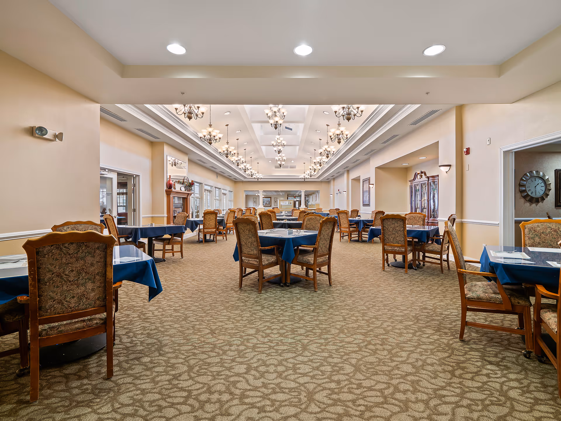 Spacious dining room with multiple tables covered in blue tablecloths and surrounded by wooden chairs with patterned upholstery. The room features beige walls, a patterned carpet, and elegant chandeliers hanging from a coffered ceiling. Large windows line one side of the room, and decorative cabinets and wall art are visible.