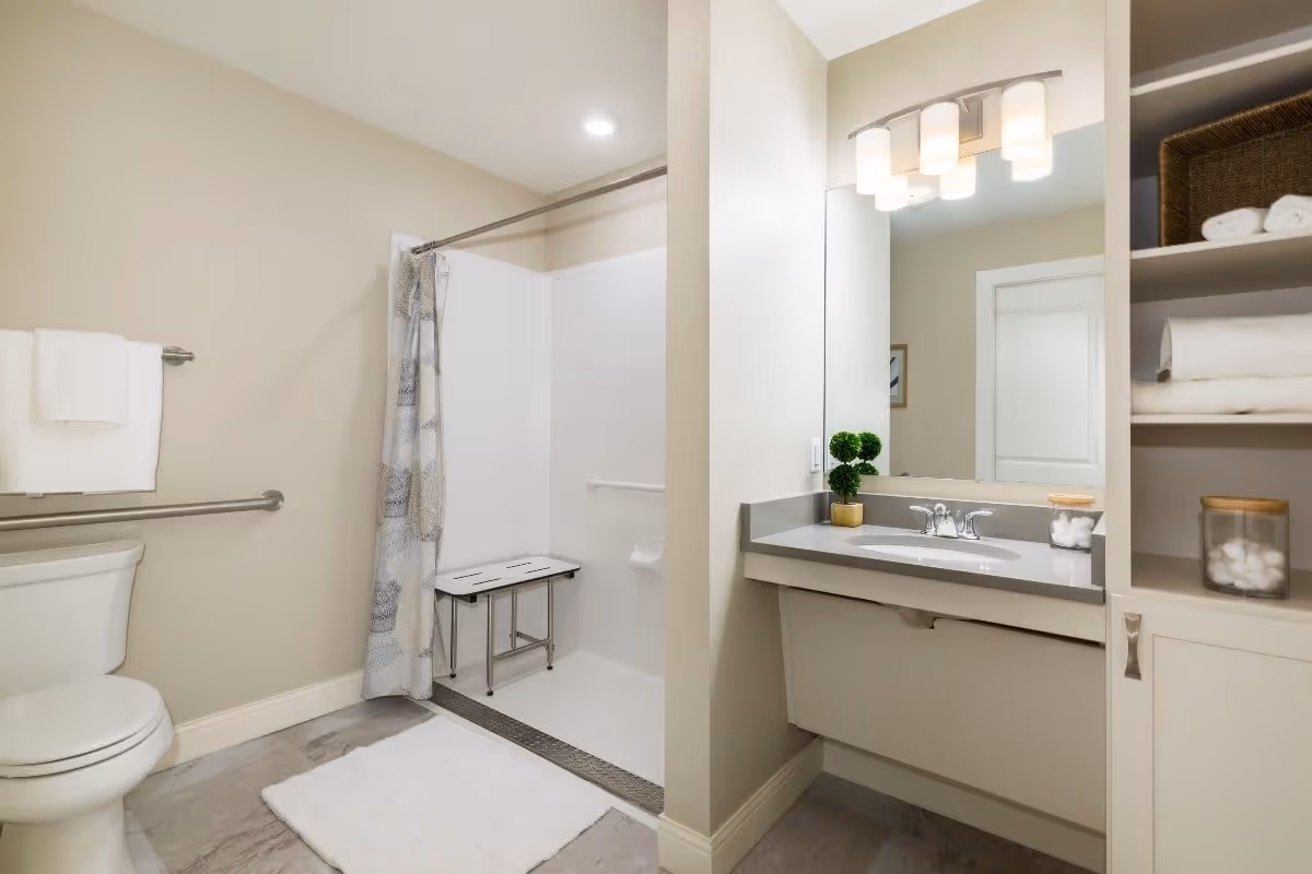 A clean and modern bathroom featuring a toilet with a towel rack above it, a walk-in shower with a bench and a patterned shower curtain, a vanity with a sink, a large mirror, and overhead lighting. Shelves next to the vanity hold neatly folded towels and jars with cotton balls.