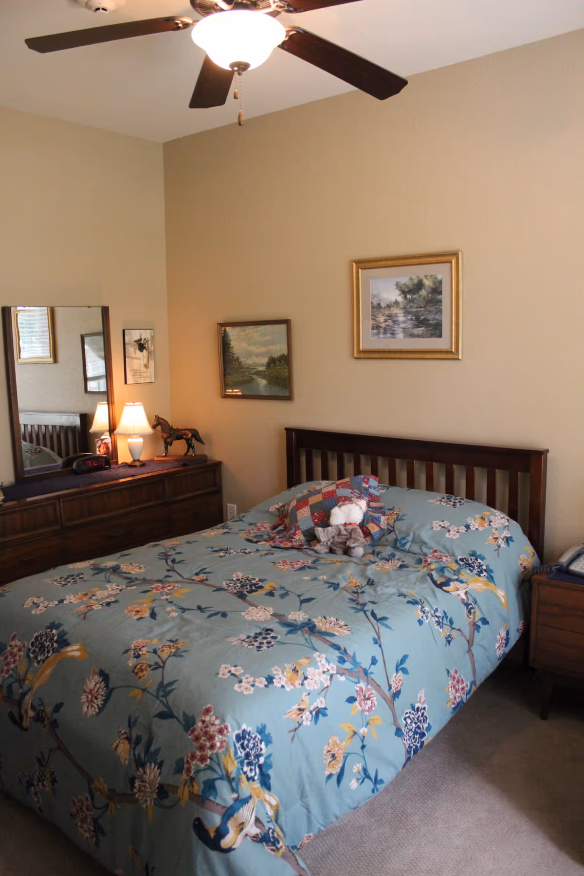 A cozy bedroom with a floral bedspread on a wooden bed, dresser with mirror and lamp, framed wall art, and a ceiling fan.