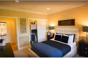 A cozy bedroom with a neatly made bed featuring white and dark blue bedding, two black nightstands with lamps on either side, a decorative wall hanging above the bed, and a closet with open doors showing clothes inside. The walls are painted a warm beige color and the room is well-lit with recessed ceiling lights.