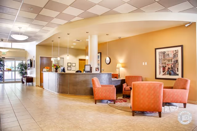 A bright and welcoming reception area in a senior living facility with a curved front desk, four orange upholstered armchairs arranged around a circular rug, a table lamp, framed artwork on the wall, and large windows letting in natural light.