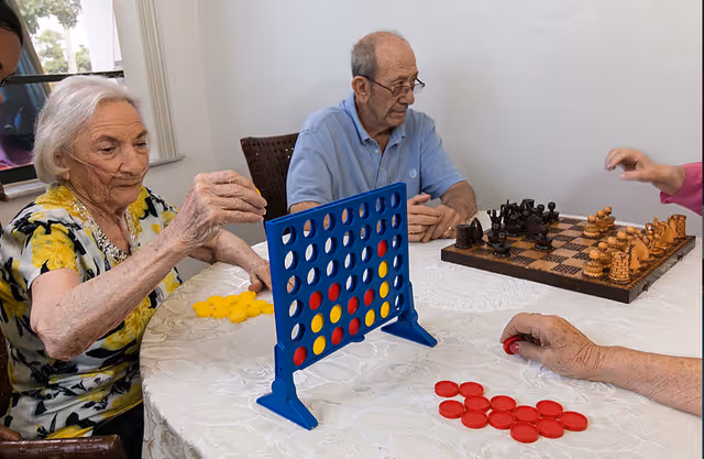 Three elderly individuals sitting around a table playing board games. One woman with white hair and a floral shirt is placing a yellow disc in a blue Connect Four game. A man in a light blue polo shirt is sitting next to a chessboard with chess pieces arranged for a game. Another person’s hand is visible holding a red disc near the Connect Four game. The table is covered with a white lace tablecloth.