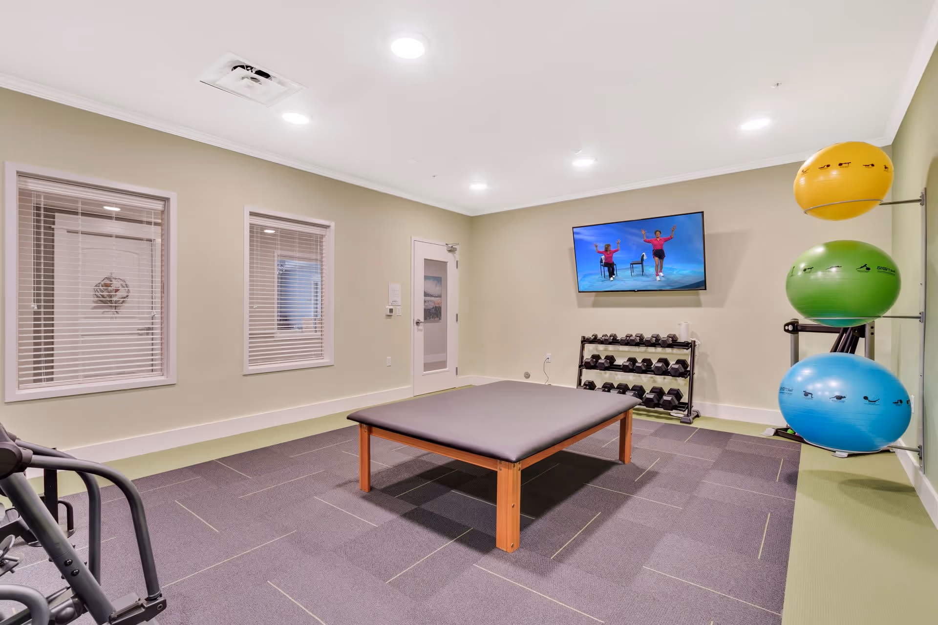 A fitness room with a large padded exercise table in the center, a rack of dumbbells against the wall, three large exercise balls mounted on a rack, and a wall-mounted TV displaying an exercise video with two people performing seated exercises. The room has light green walls, two windows with blinds, and recessed ceiling lights.