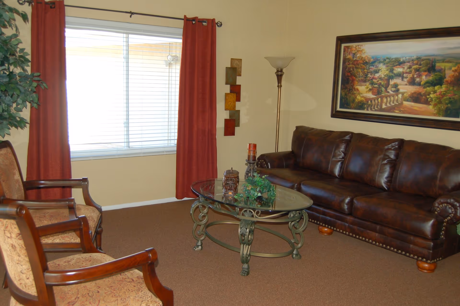A cozy living room with a brown leather sofa, glass coffee table, two upholstered chairs, and a window with red curtains.