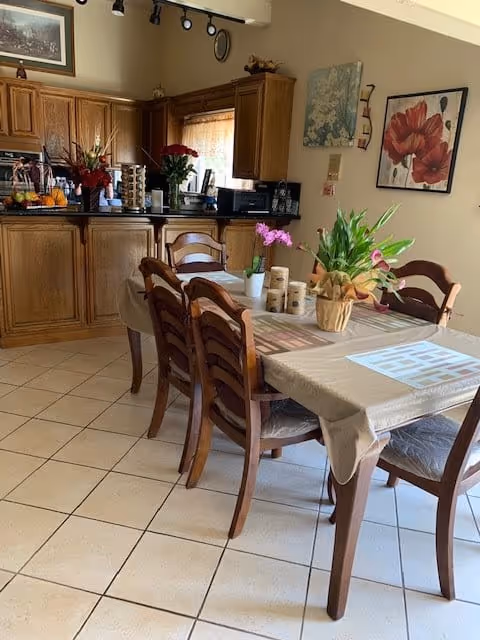 A dining area with a wooden table covered by a beige tablecloth and six wooden chairs. The table has potted plants and decorative items on it. In the background, there is a kitchen with wooden cabinets, a countertop with various items, and a window with a curtain. The walls have framed floral artwork and a clock.