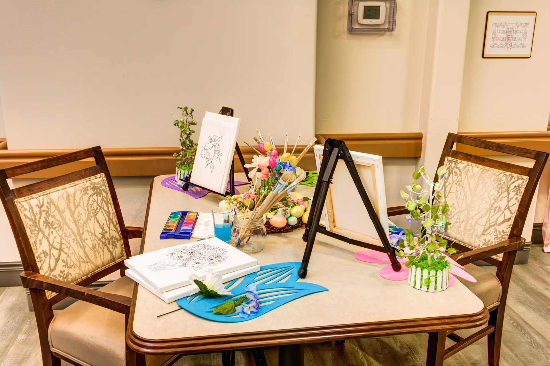 A table set up for an arts and crafts activity with two wooden chairs. On the table are coloring pages, paintbrushes in a jar, watercolor paints, decorative flowers, and small easels holding blank canvases. The room has light-colored walls and wood flooring.