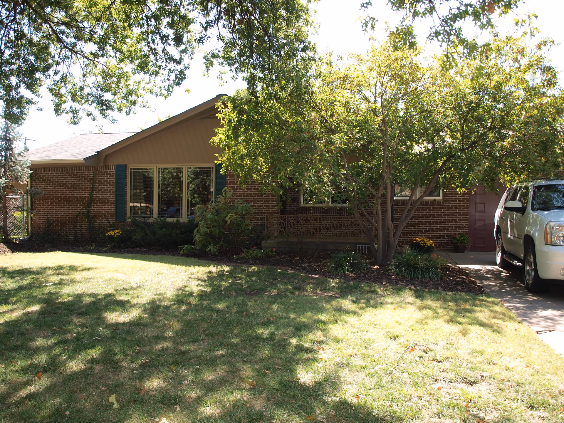 Front exterior view of a single-story brick house with a sloped roof, surrounded by trees and a grassy lawn. A white SUV is parked in the driveway on the right side of the house.