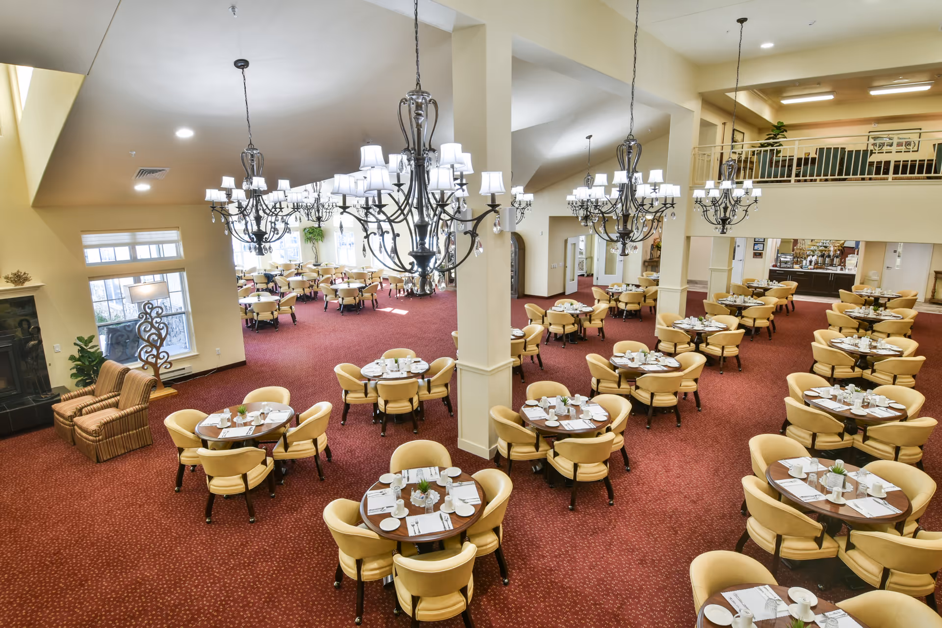 Spacious dining room with multiple round tables set with white plates, cups, and silverware. The room features red carpet flooring, beige walls, large chandeliers hanging from the ceiling, and a seating area with two striped armchairs near a window. There is a balcony area overlooking the dining space and a serving counter in the background.