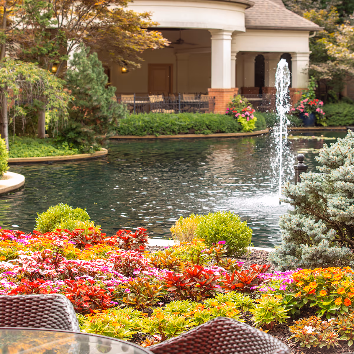 Colorful flower beds and wicker chairs beside a pond with a fountain and a covered patio in the background.