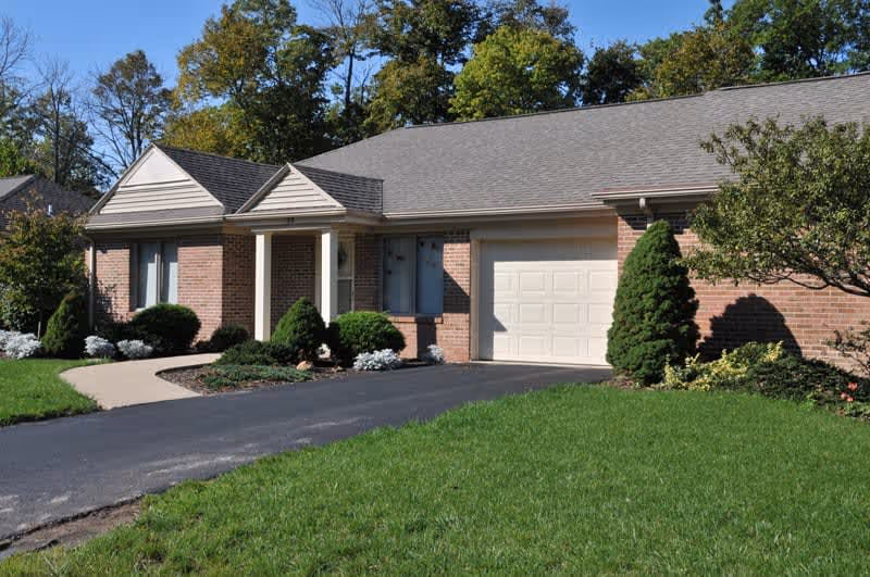 Single-story brick house with a gray shingled roof, a white garage door, and a small covered porch supported by white columns. The house is surrounded by neatly trimmed bushes, a green lawn, and trees in the background under a clear blue sky.