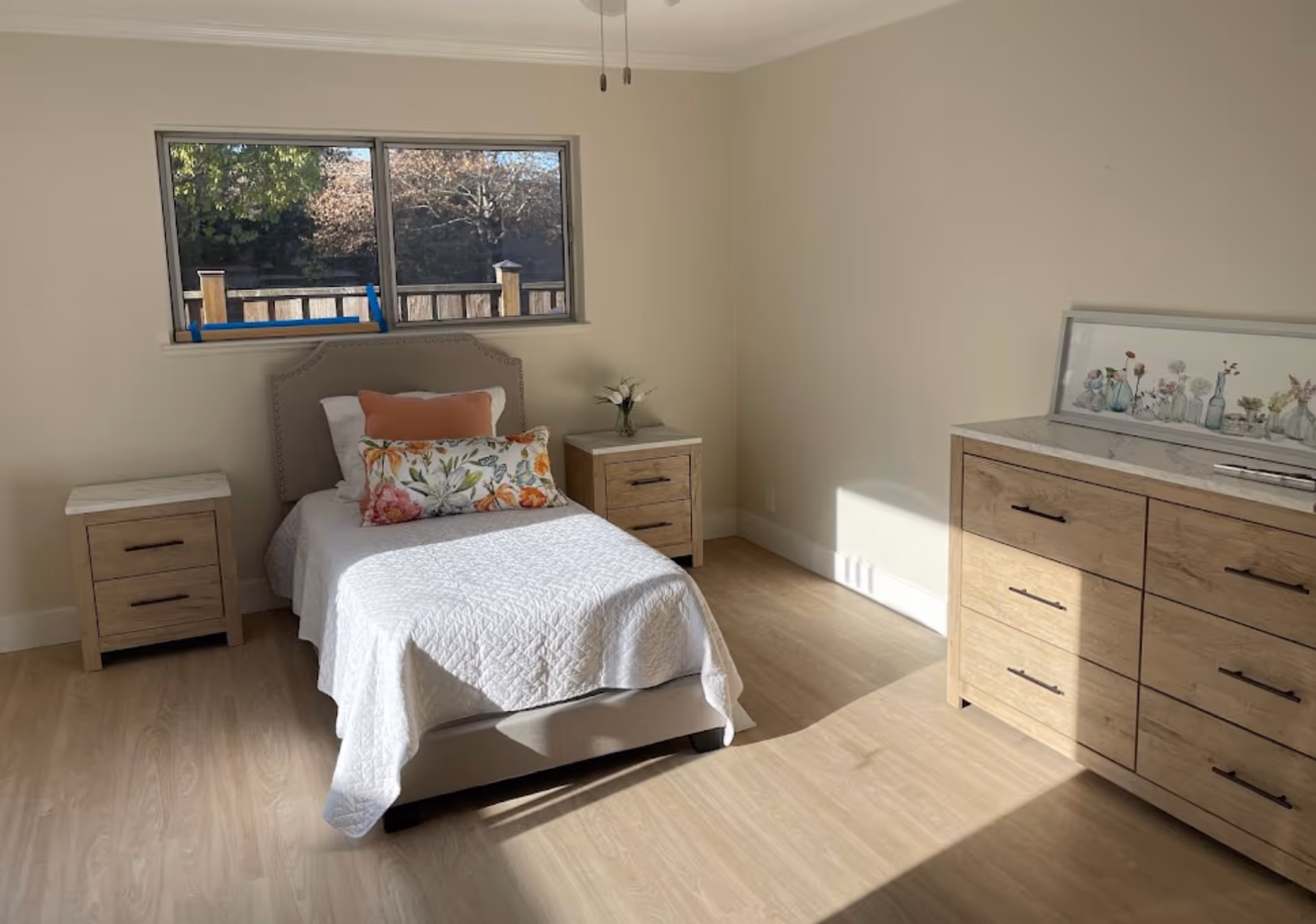 Sunlit bedroom with a single bed, two nightstands and a dresser beneath a window.