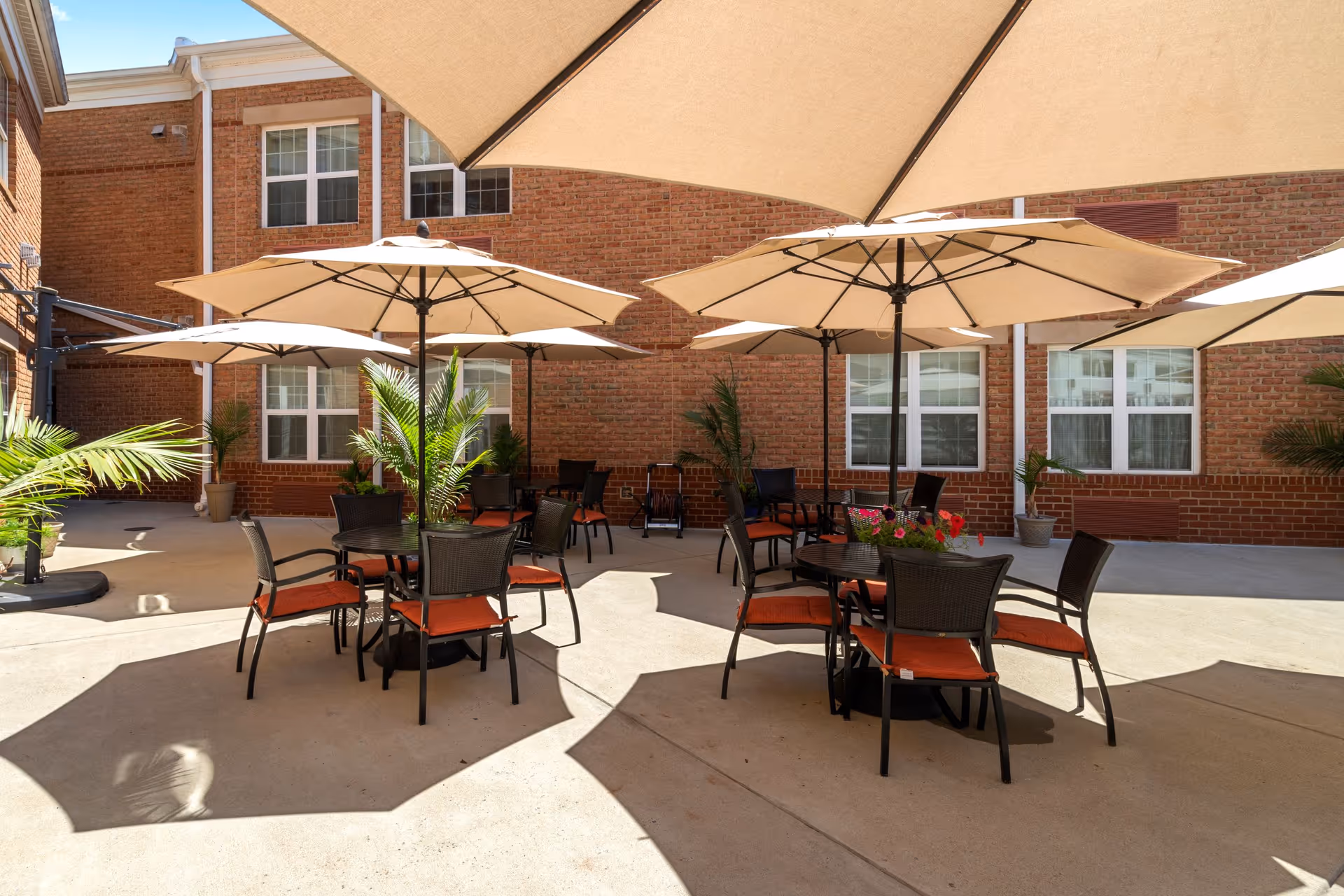 Outdoor patio area with multiple round tables and chairs, each table shaded by large beige umbrellas. The patio is surrounded by a brick building with several windows, and there are potted plants placed around the space.