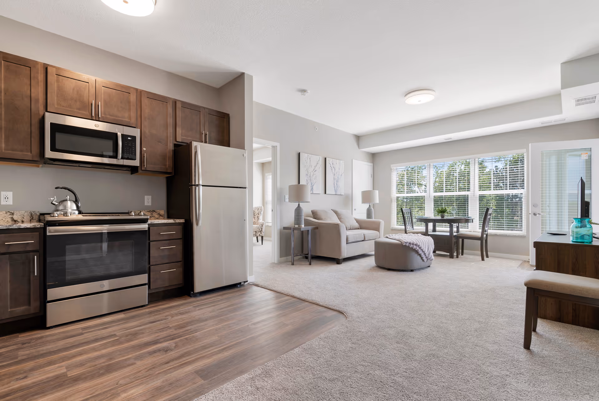 Open-concept senior living apartment with a kitchen area featuring stainless steel appliances including a microwave, stove, and refrigerator, dark wood cabinets, and wood flooring. Adjacent living room area has a beige sofa, two side tables with lamps, a round ottoman with a throw blanket, a small dining table with chairs near large windows letting in natural light, and a TV on a stand.