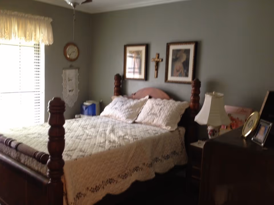 A cozy bedroom with a wooden bed frame featuring turned posts, a white quilted bedspread, and two pillows. The room has a window with blinds and a valance on the left side, a clock and a hanging decorative piece on the wall, two framed pictures and a crucifix above the bed. A nightstand with a lamp and some framed photos is on the right side of the bed.