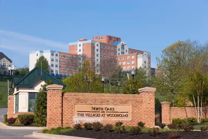 Entrance to North Oaks and The Villages at Woodholme facility with a brick sign in the foreground and a large multi-story building in the background surrounded by trees and greenery under a clear blue sky.