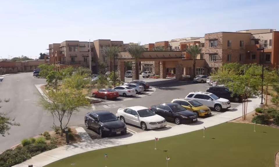 Exterior view of The Palms At La Quinta Gracious Retirement Living facility showing a parking lot with several cars, a putting green in the foreground, and a multi-story building with balconies and palm trees in the background under a clear sky.