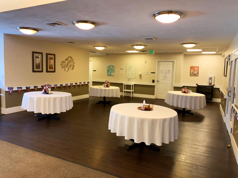 Interior view of a dining room with four round tables covered with white tablecloths, each set with condiments and napkins. The room has wooden flooring, beige walls with framed artwork, ceiling lights, and patriotic bunting along the walls.