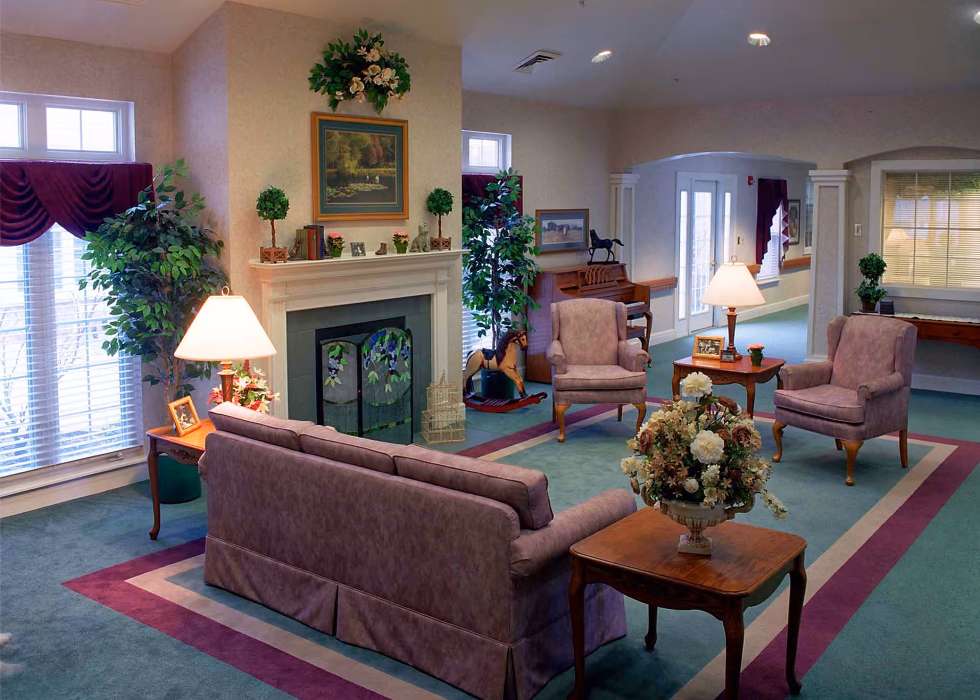A cozy living room with a purple sofa and two matching armchairs arranged around a wooden coffee table with a floral centerpiece. There is a fireplace with decorative items on the mantel, including small plants and a framed painting above it. Large windows with burgundy curtains allow natural light to fill the room. Additional furniture includes side tables with lamps and framed photos, a piano, and decorative plants.