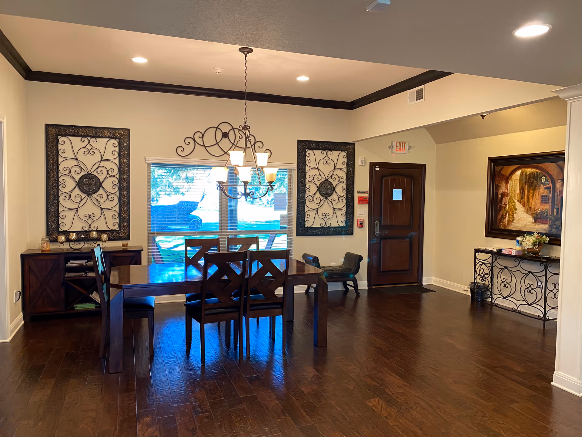 Well-lit dining area with a wooden table and chairs under a chandelier, decorative wall art, and an entry door.