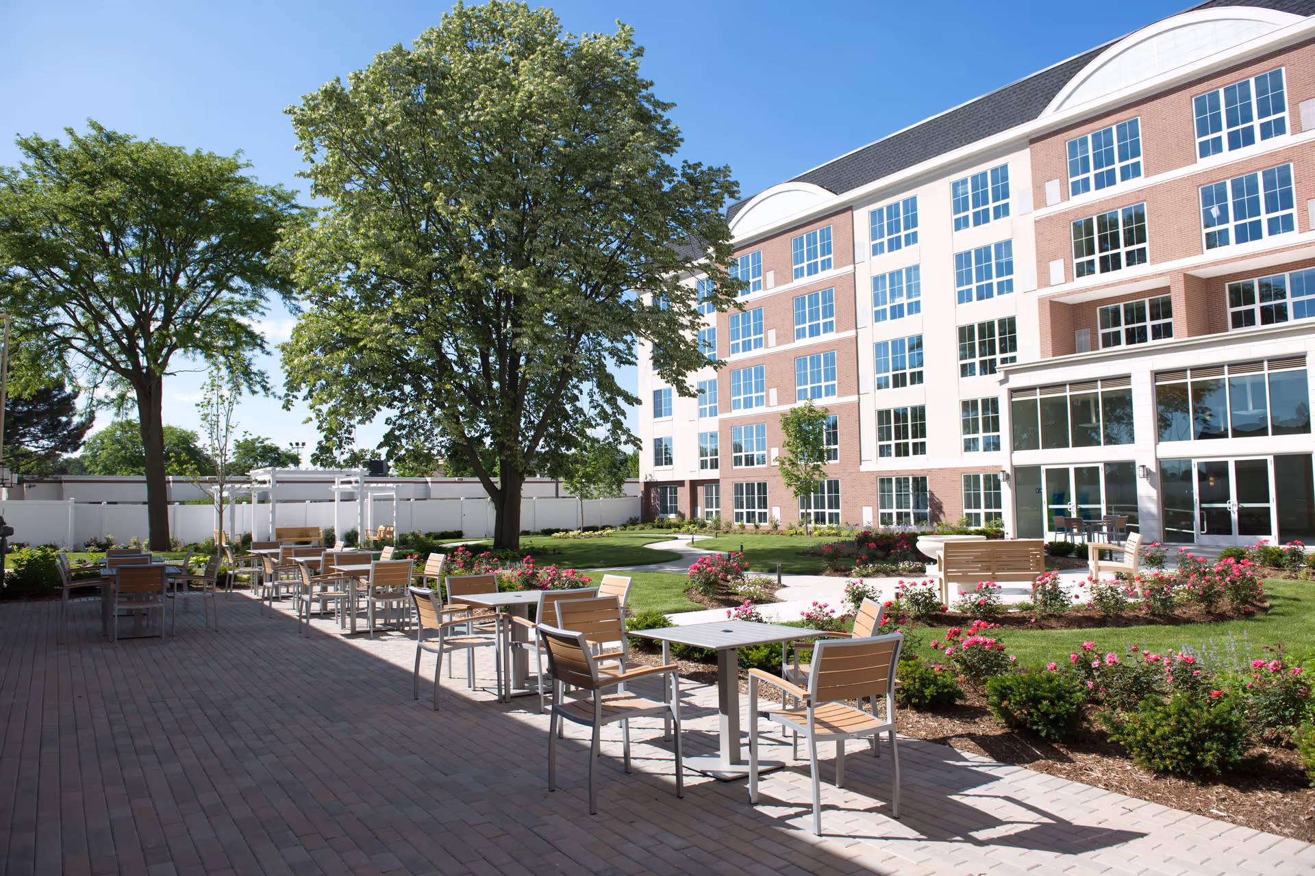 Outdoor patio area with tables and chairs surrounded by landscaped garden beds with flowers and trees, adjacent to a multi-story senior living facility building under a clear blue sky.