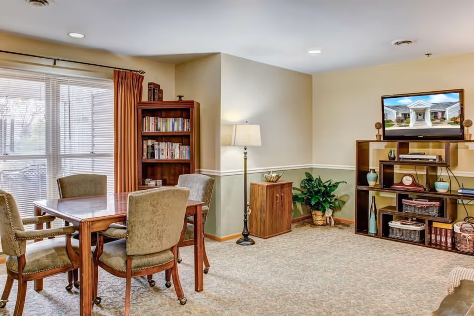 A cozy common area with a wooden table and four upholstered chairs near a large window with blinds and rust-colored curtains. A wooden bookshelf filled with books stands in the corner. A floor lamp and a small wooden cabinet are against the wall. On the right side, a wooden shelving unit holds decorative items and a flat-screen TV displaying an image of a building entrance. The room has beige walls with a green lower half and carpeted floor.
