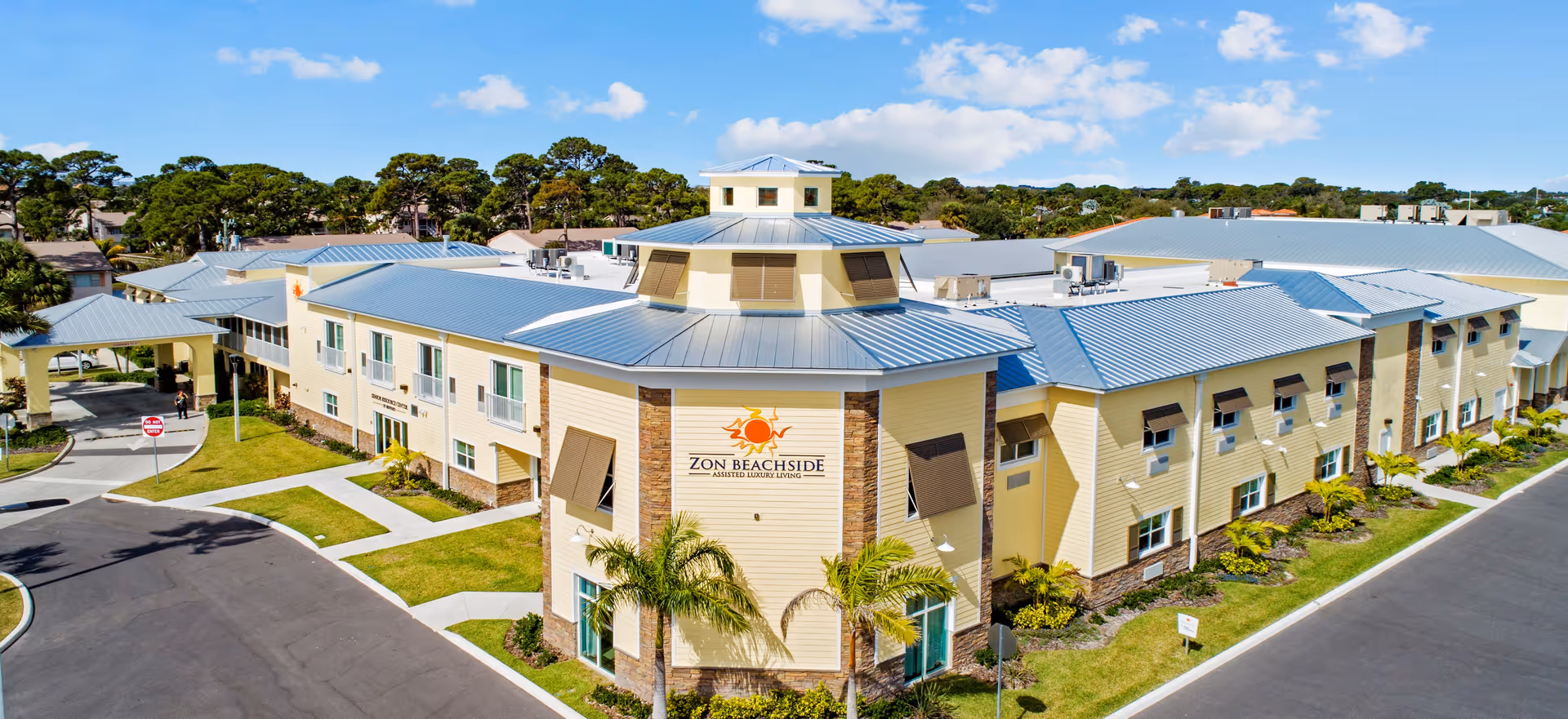 Aerial view of Zon Beachside, a large assisted living facility with light yellow exterior walls and a metal roof. The building is surrounded by well-maintained landscaping with palm trees and green lawns under a bright blue sky with scattered clouds.