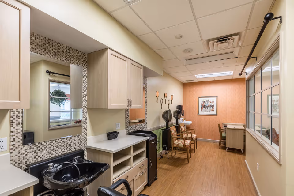 Interior view of a salon area in a senior living facility featuring a black shampoo sink with a chair, light wood cabinets, a mosaic tile backsplash, two salon hair dryers with chairs, a small desk with a chair, and framed artwork on the wall. The floor is wood, and there is a large window on the right side.